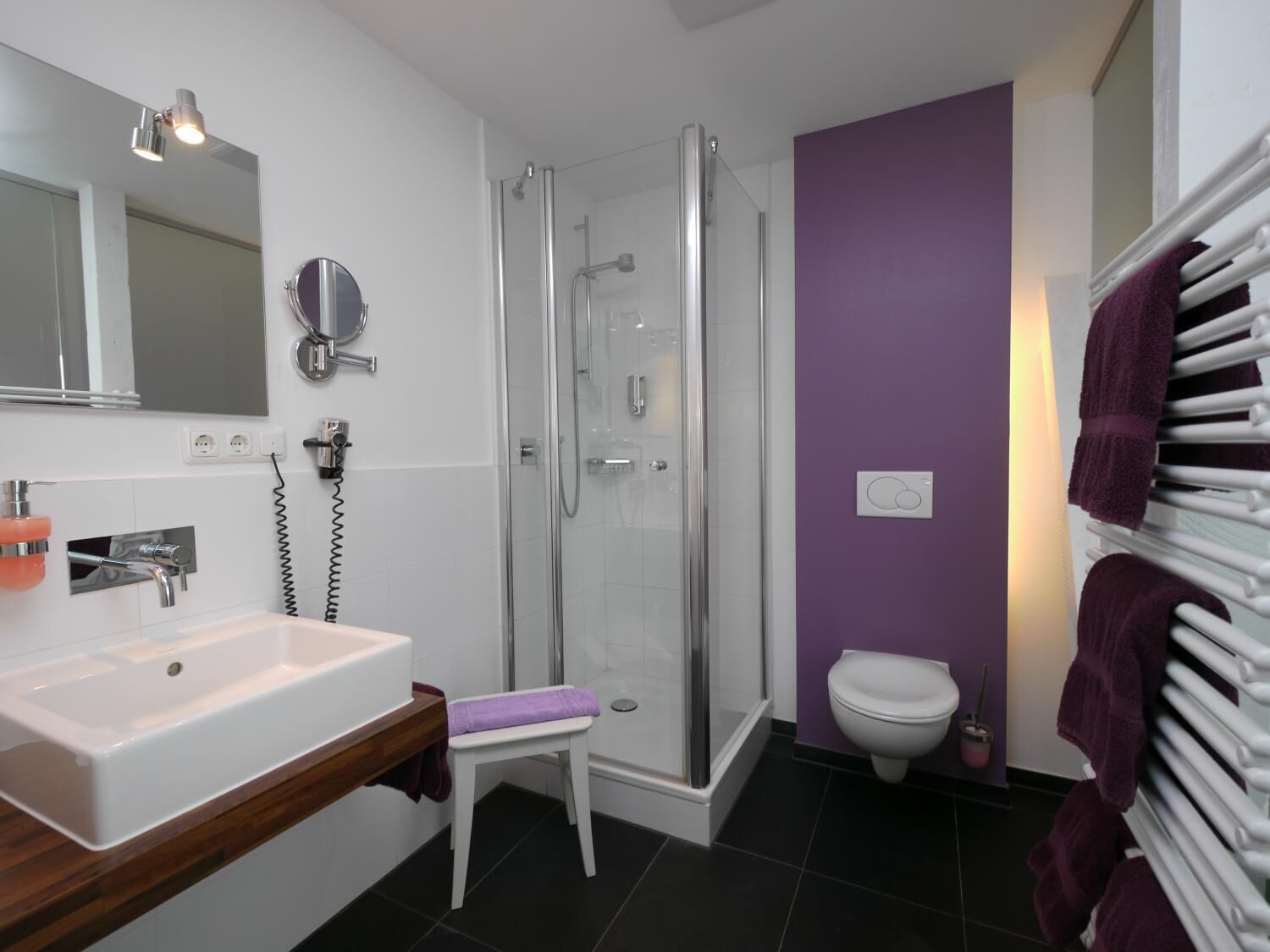 Un baño moderno con ducha envuelta en vidrio, un lavabo blanco y una pared morada.