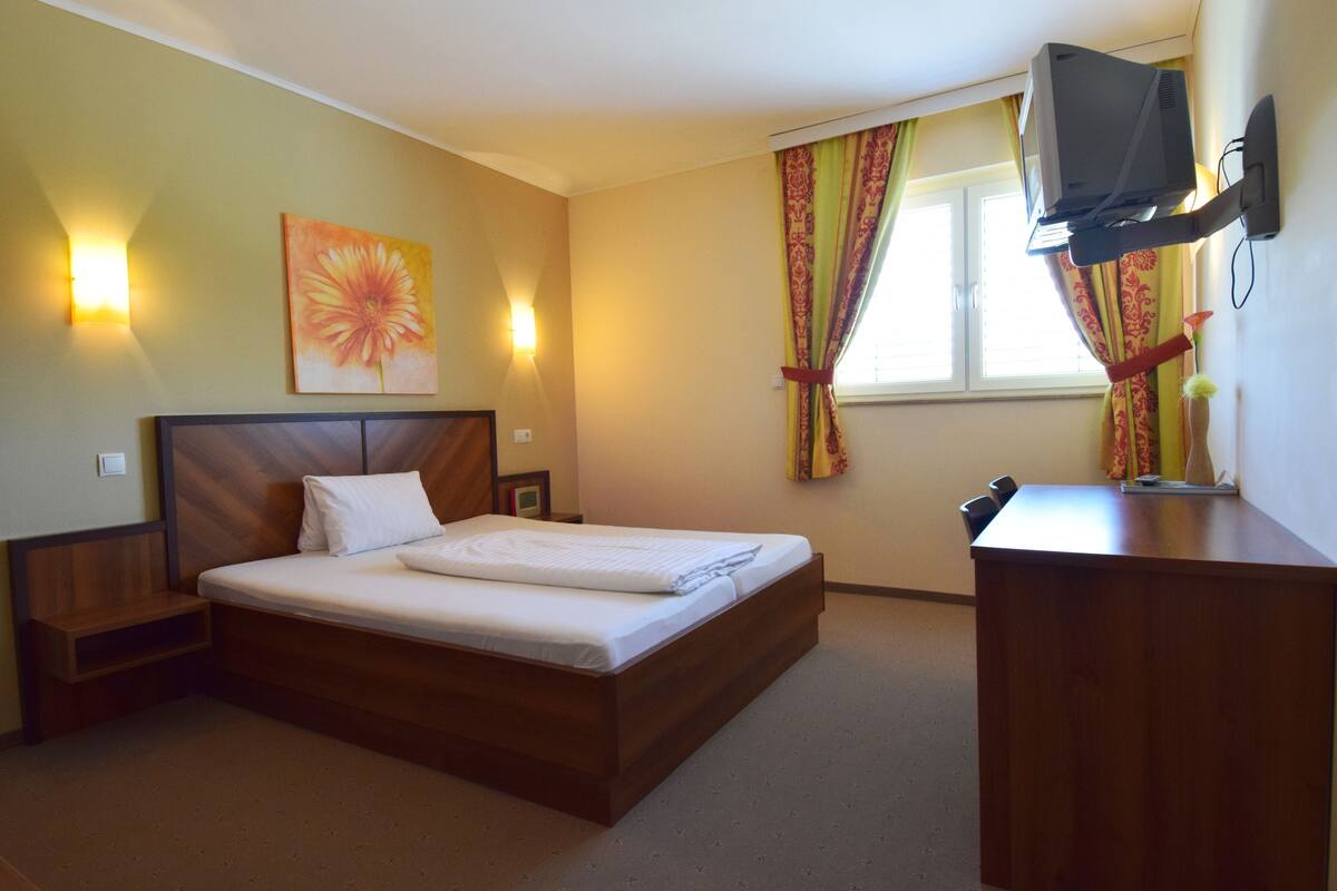 Quarto de hotel com cama de madeira, escrivaninha, televisão e janela com cortinas.