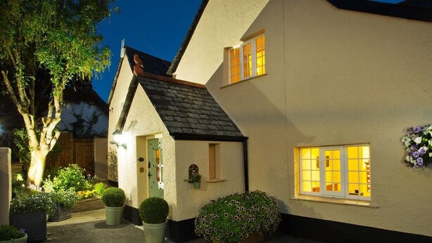 A quaint, well-lit cottage with a sloped roof, surrounded by potted plants and a small garden. - Tinhay Mill Guest House (Lifton)