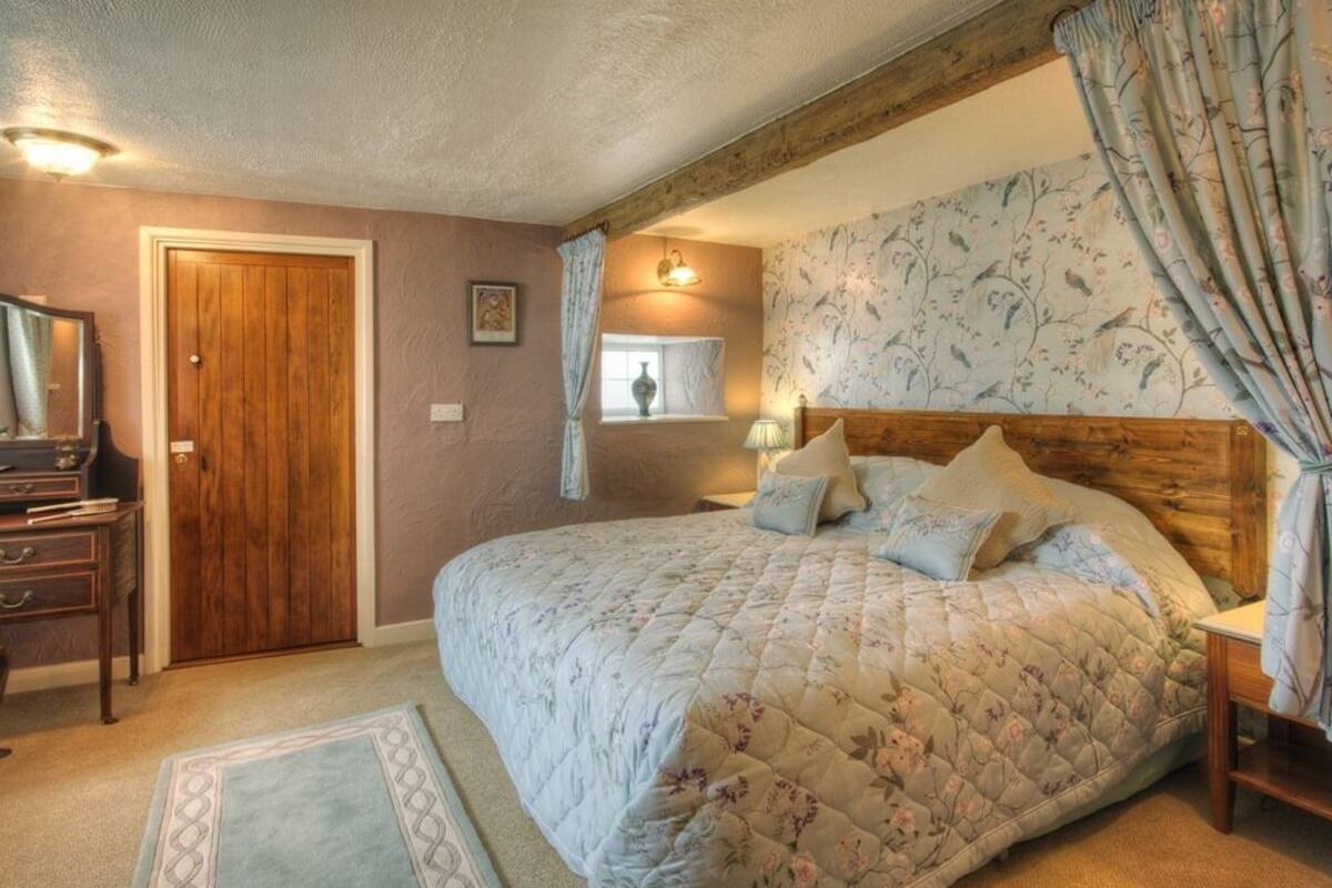 Un dormitorio con cabecera de madera, edredón estampado con motivos florales, mesita de noche con lámpara y ventana con cortinas.