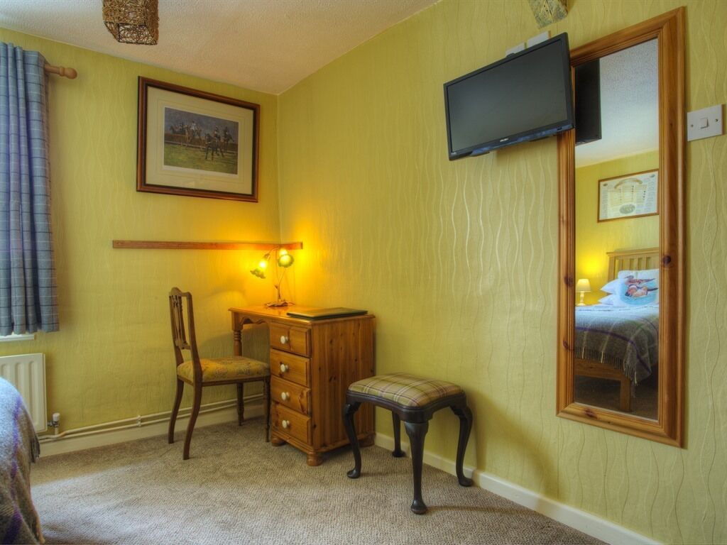A room with a wooden dresser, a chair, a small table with a lamp, a television mounted on the wall, and a bed with a headboard.