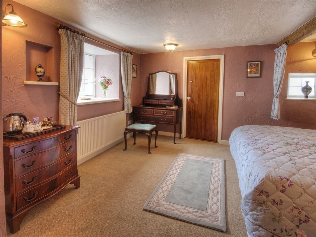 A bedroom with a wooden dresser, a vanity, a bed with a floral quilt, a window with curtains, and a radiator.