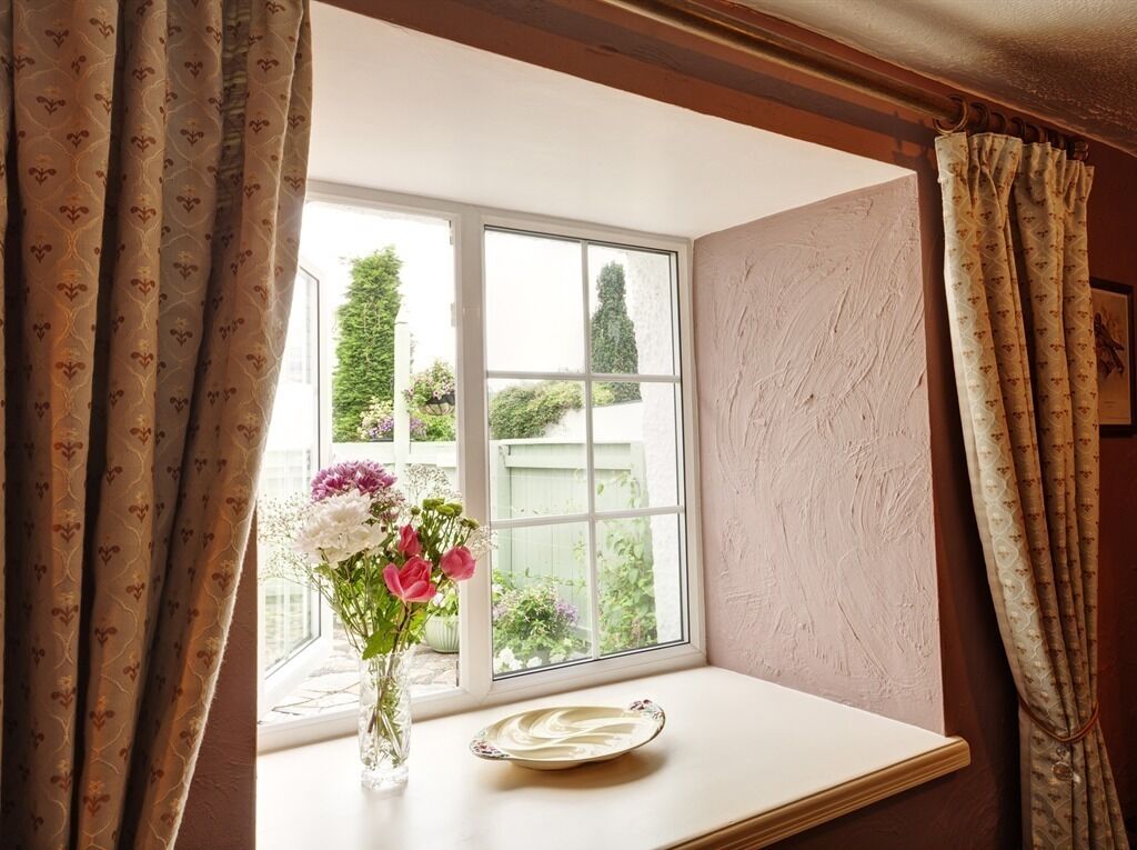 A window with a view of greenery and a white table with a vase of flowers and a plate.