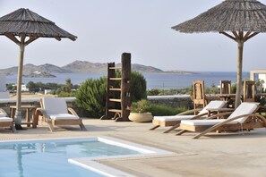 A poolside area with lounge chairs and umbrellas, overlooking a body of water and hills. - Anemoi Resort (Paros)