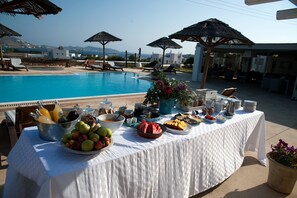 A poolside buffet with a variety of fruits and beverages, surrounded by lounge chairs and umbrellas. - Anemoi Resort (Paros)