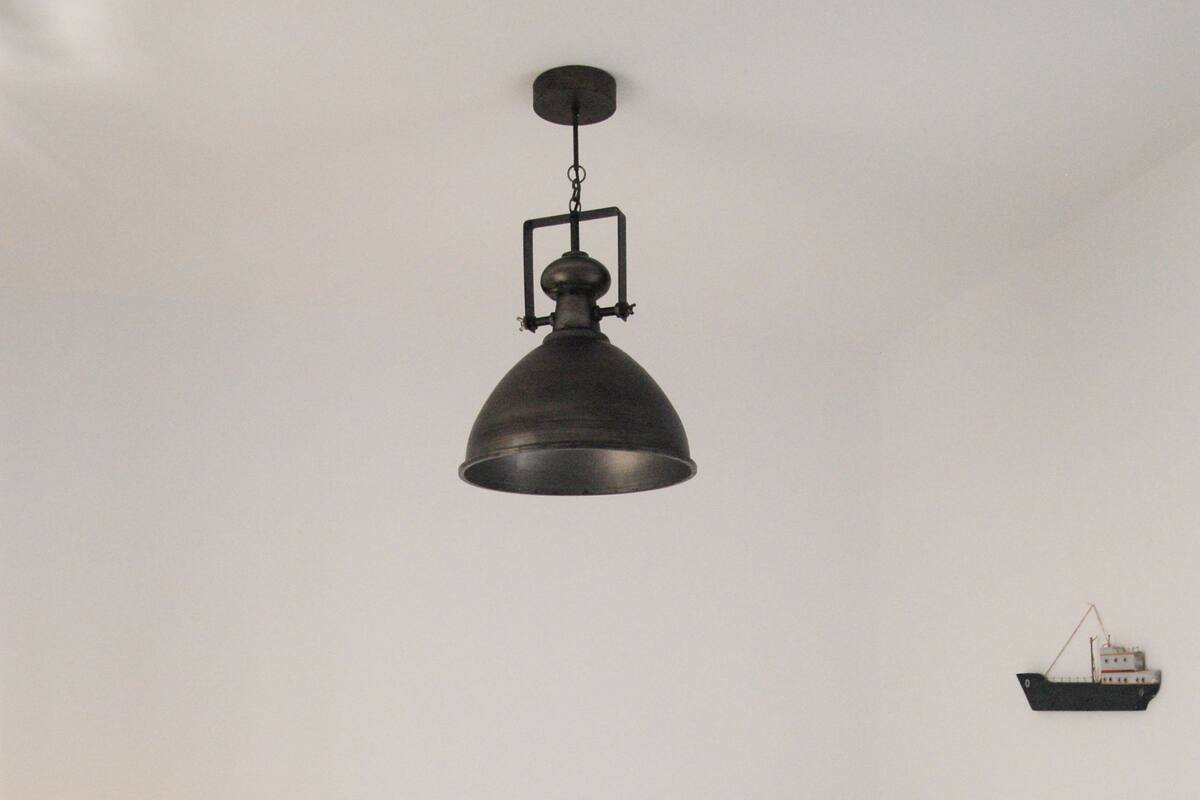 Una cama bien hecha con sábanas blancas y una flor roja, una lámpara colgante negra y una pequeña decoración de barco en la pared.