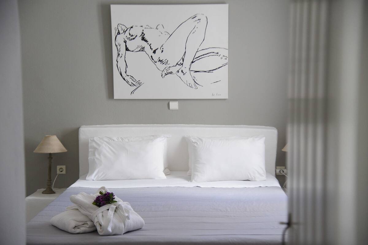 Una habitación de hotel con cama, ropa de cama blanca, una toalla con un detalle floral, una lámpara y una obra de arte en la pared.