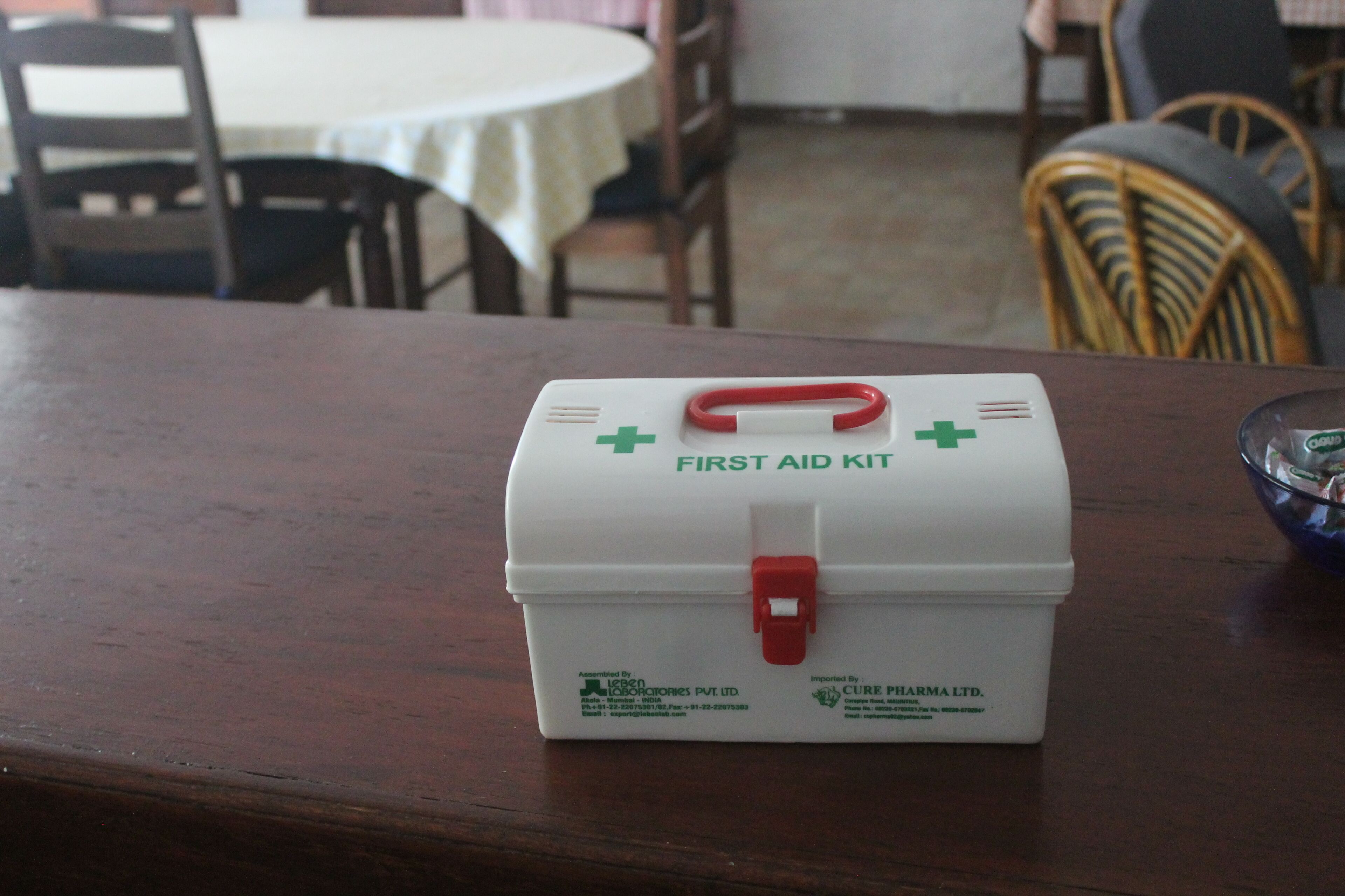 Une trousse de premiers secours blanche avec une poignée rouge, posée sur une table en bois dans un coin repas.