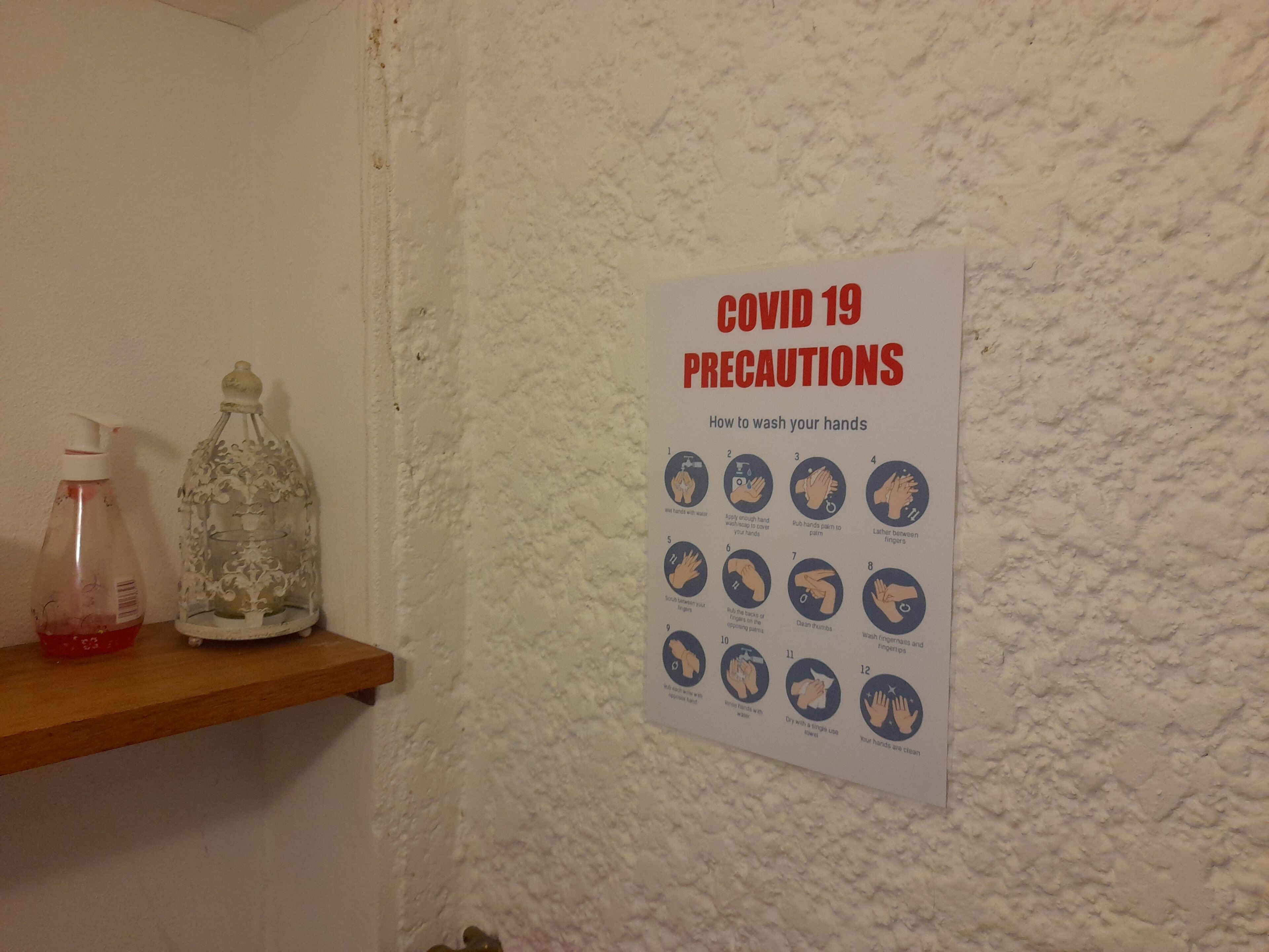 Un cartel sobre precauciones por COVID-19 en una pared texturizada, una estantería de madera con un pulverizador y un recipiente de vidrio decorativo.