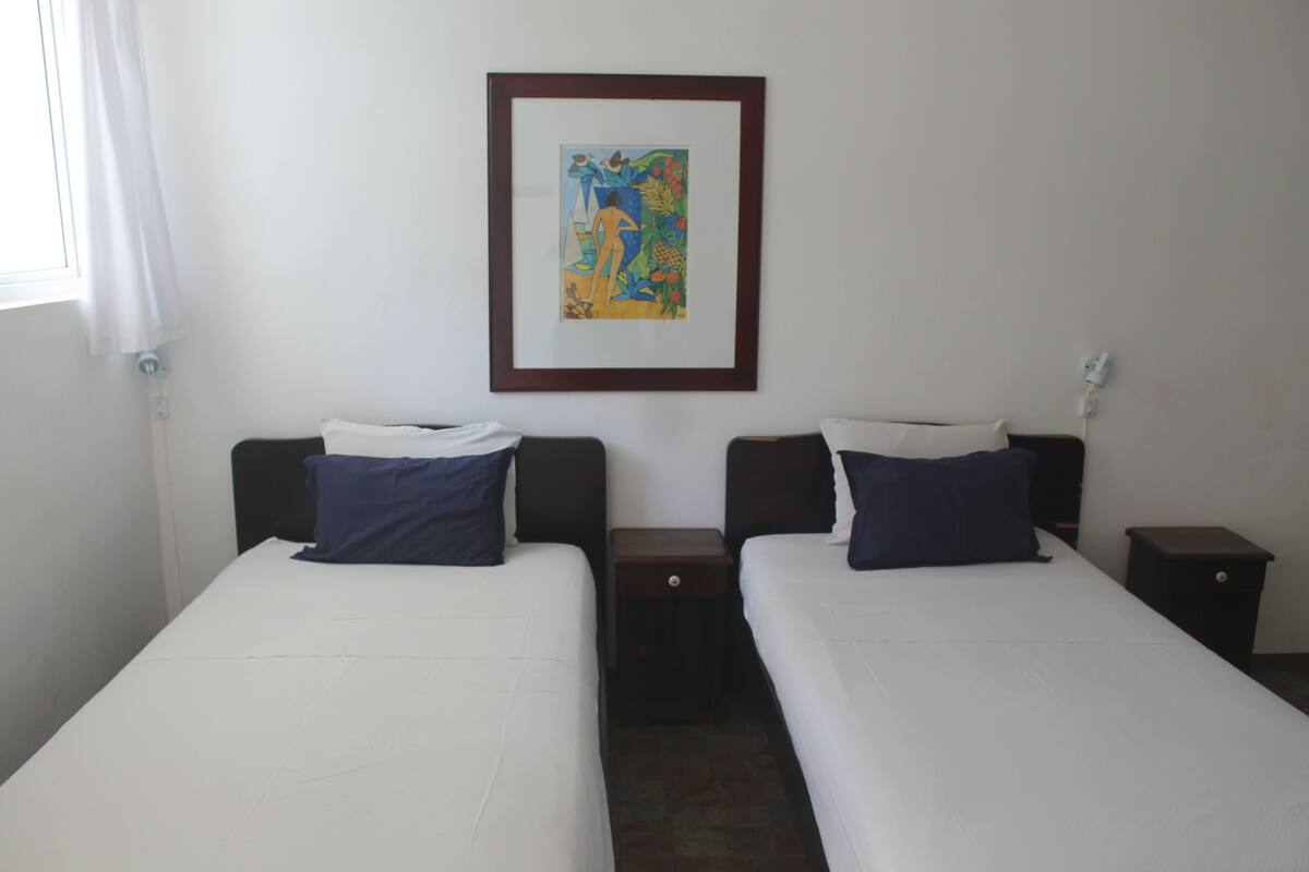 A hotel room with two single beds, a nightstand with a lamp, a framed artwork on the wall, and a window with a white curtain.