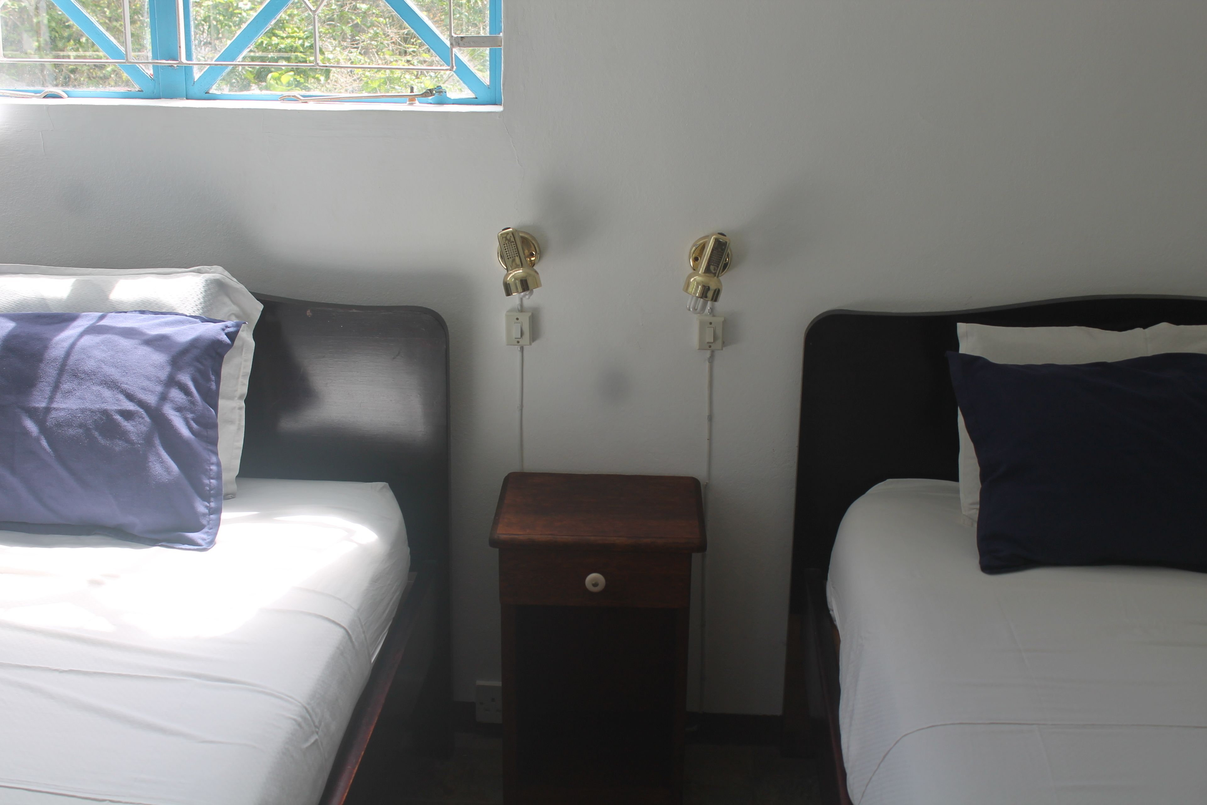 A room with two beds, each with a pillow and a nightstand with a drawer. There are two wall-mounted lamps.