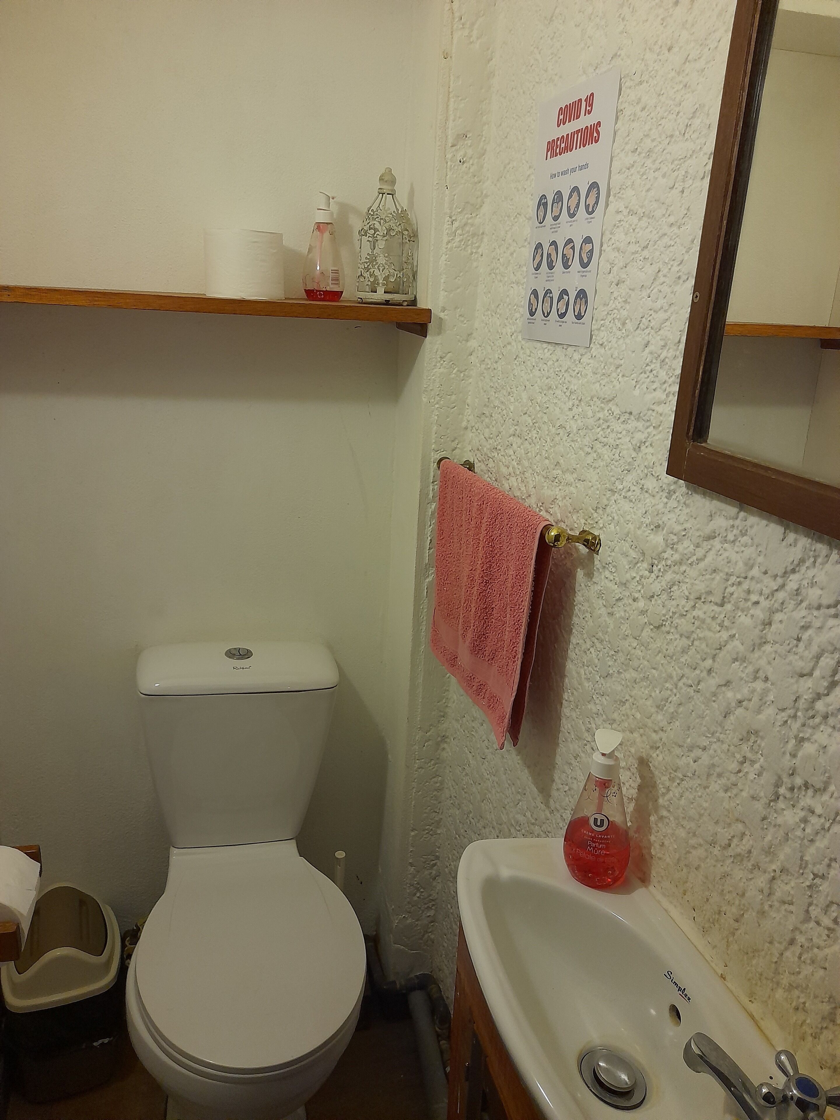 A bathroom with a toilet, sink, and a hand sanitizer dispenser. There is a 'COVID 19 PRECAUTIONS' sign on the wall.