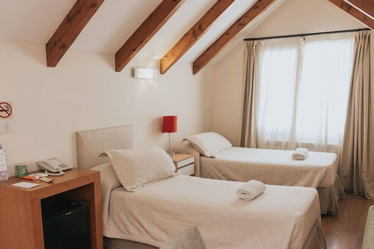 Quarto de hotel com duas camas, uma escrivaninha com abajur, telefone e janela com cortinas.