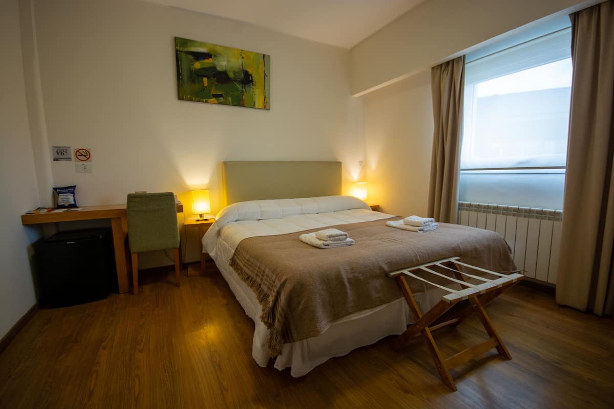 Quarto de hotel com cama, mesa de cabeceira, luminária e janela com cortinas.