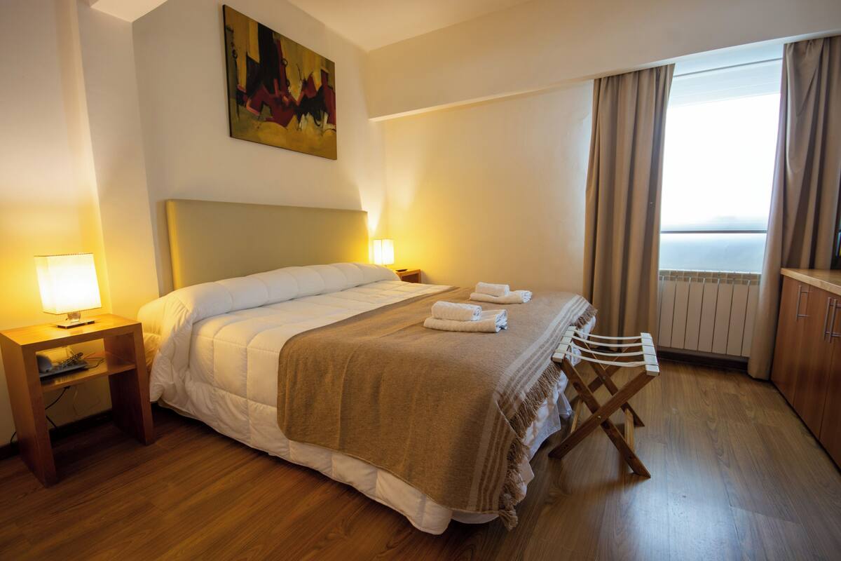 Quarto de hotel com cama, mesa de cabeceira, cadeira, pintura e vista para o mar.
