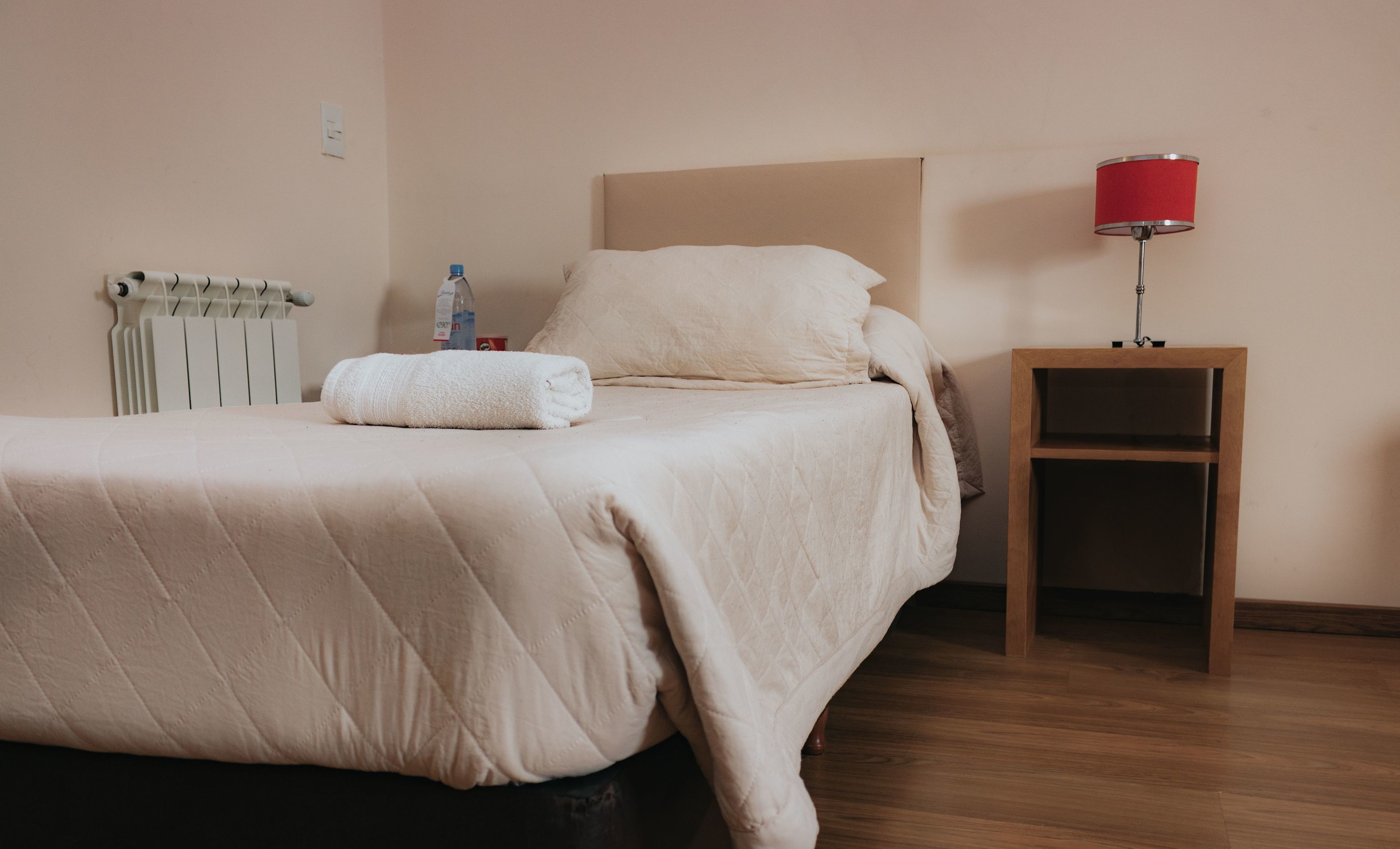 Una cama bien hecha con edredón blanco, una mesita de noche de madera con una lámpara roja, y un radiador al fondo.