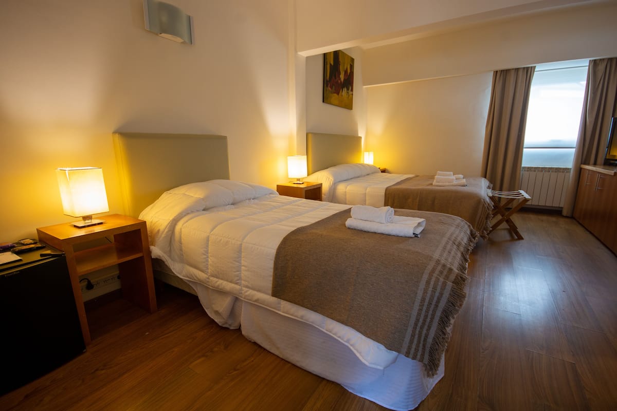 Quarto de hotel com duas camas, piso de madeira, mesas de cabeceira com abajures, um espaço de estar pequeno e vista para o mar pela janela.
