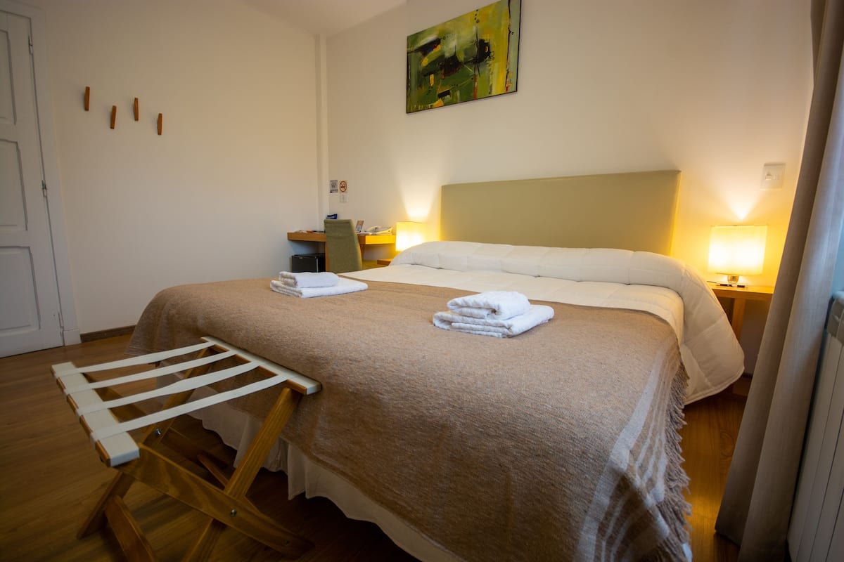 Cama bem arrumada com manta marrom, cadeira de madeira e escrivaninha com cadeira e luminária.