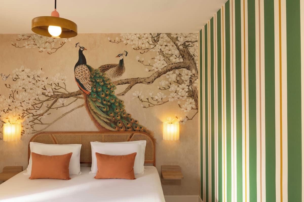 Una habitación con cama, almohadas, un mural de pavo real, apliques de pared y una pared a rayas.