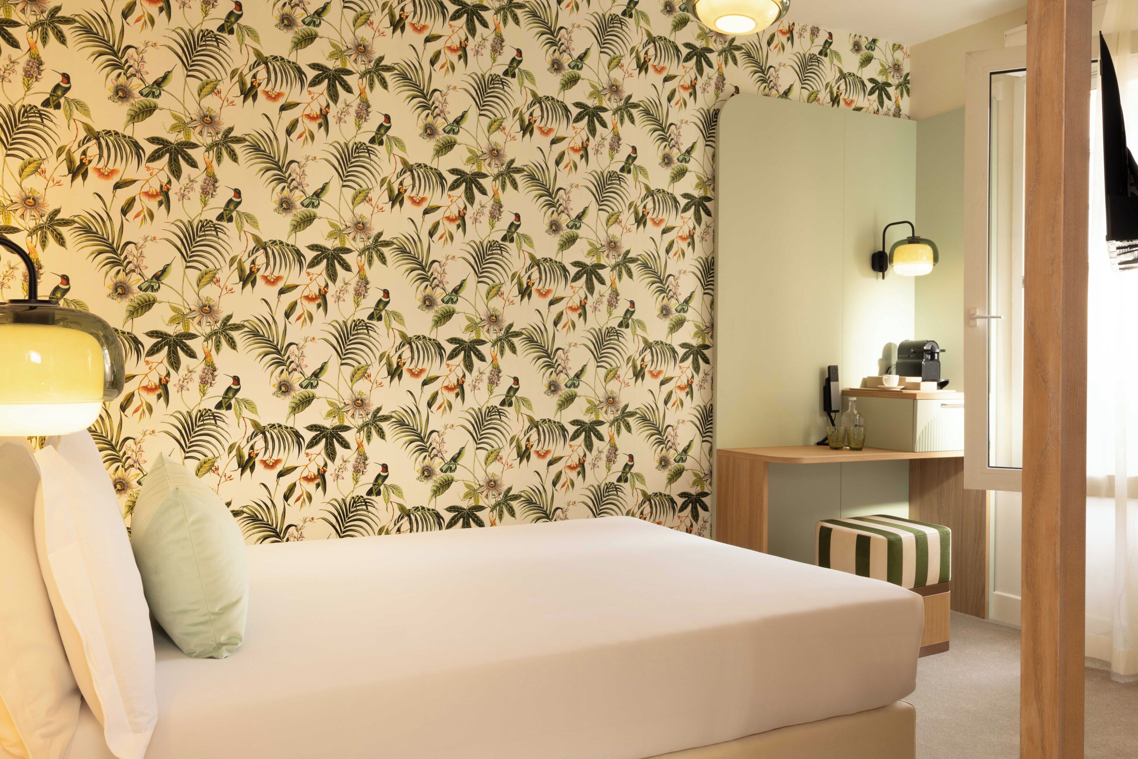 Une chambre à coucher comprenant un lit, une table de chevet, une lampe, un miroir et un mur orné d’un motif de feuilles tropicales.
