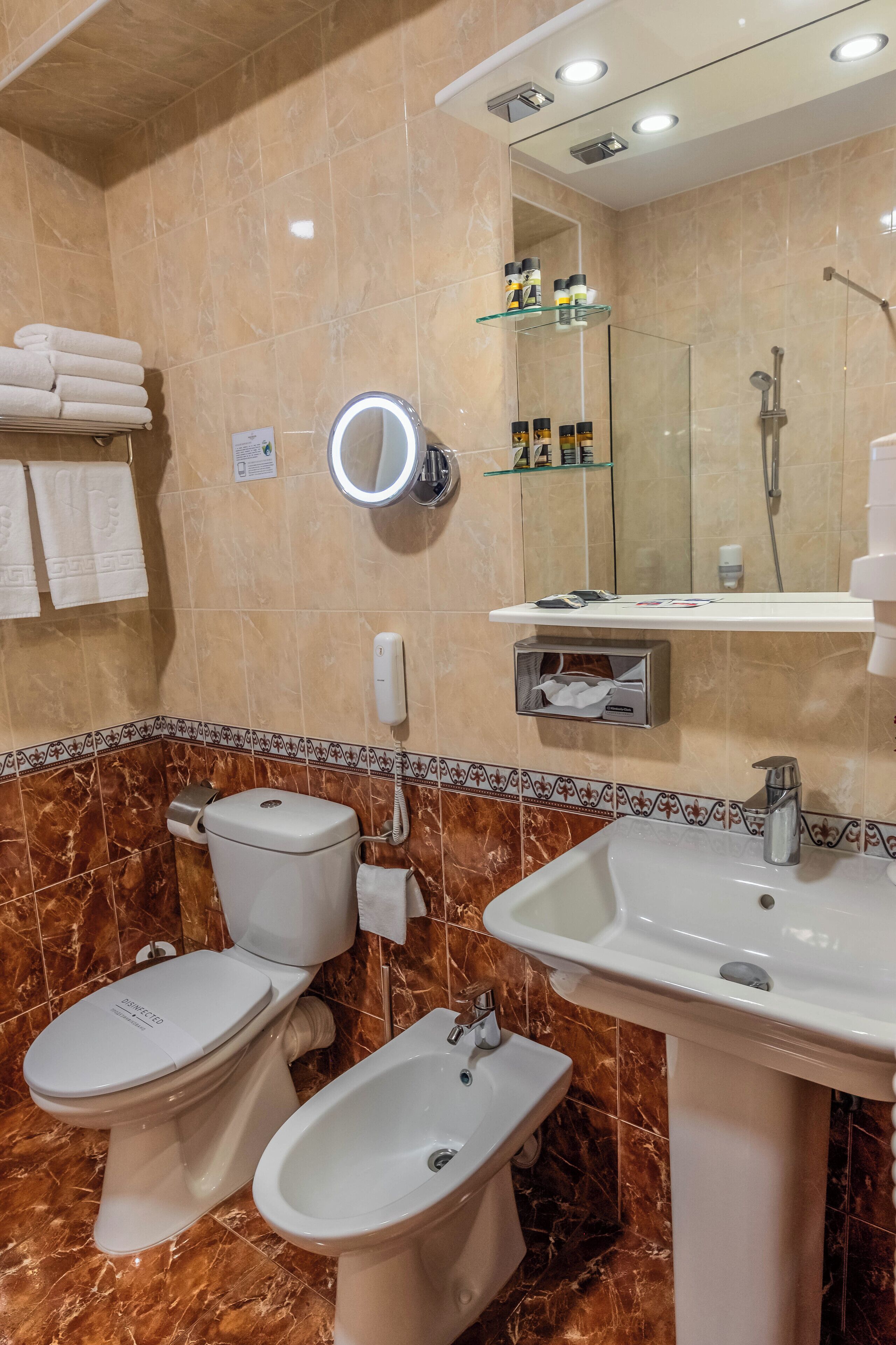 Un baño con inodoro, bidé, lavabo y espejo con iluminación. Hay toallas en un perchero y una repisa de vidrio con artículos de aseo.