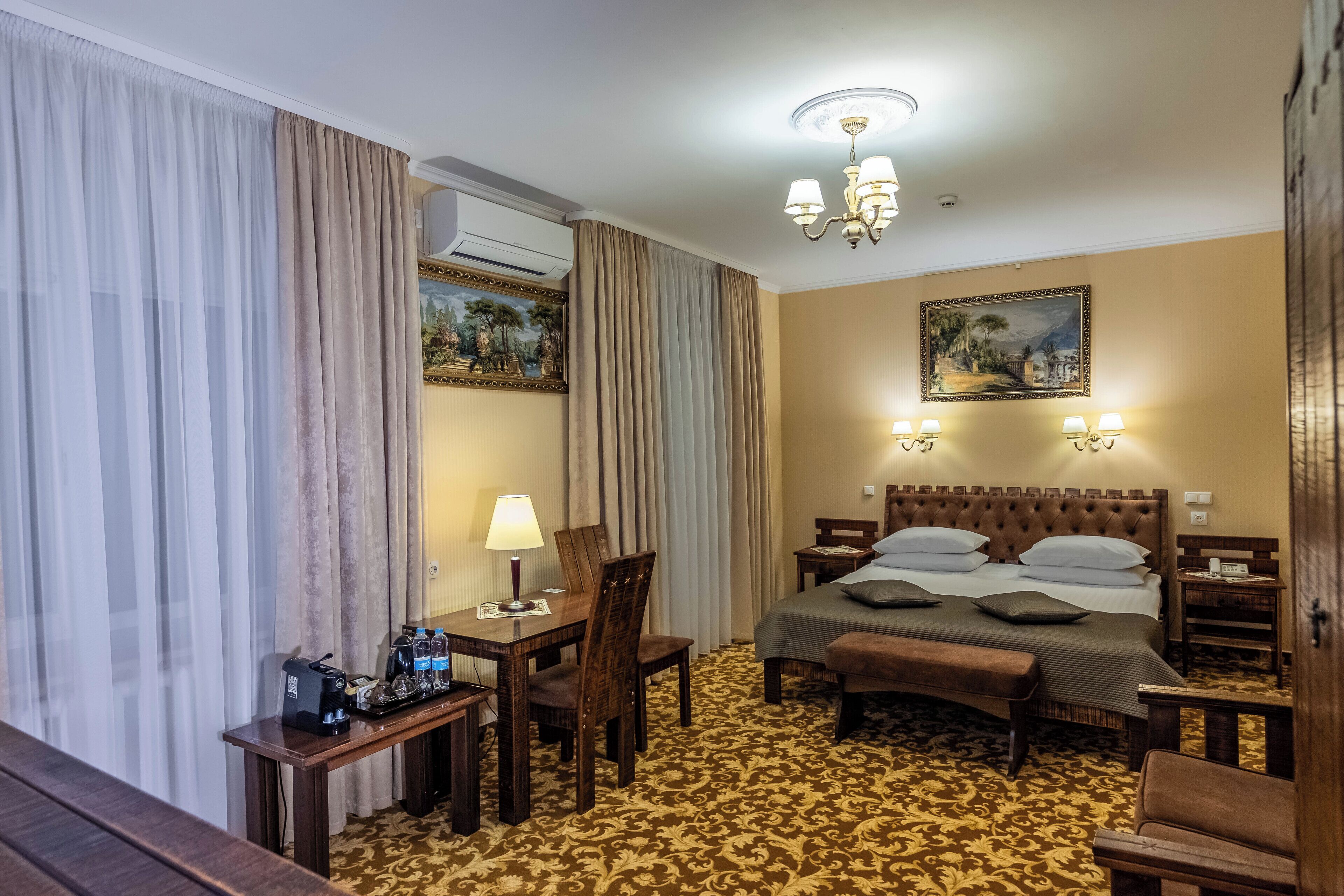 A hotel room with a bed, a desk with a lamp, a chair, a bench, and a painting on the wall.