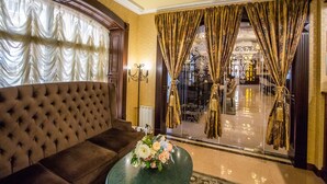 Una lujosa sala de estar con un sofá marrón capitonado, una mesa de centro de vidrio con un arreglo floral y vistas a través de una puerta de cristal con cortinas doradas.