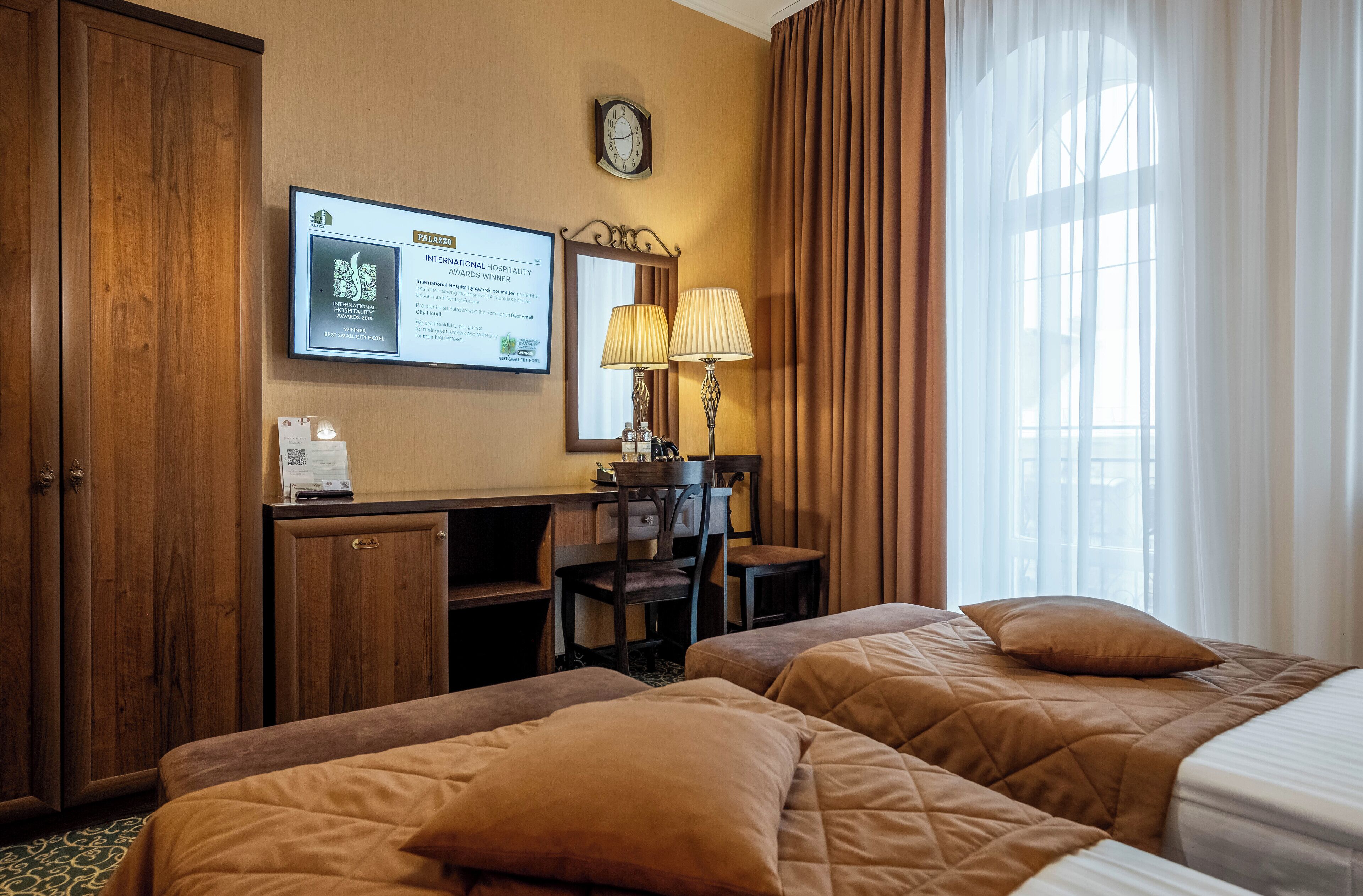 A hotel room with two beds, a desk, a chair, a lamp, and a framed award on the wall.