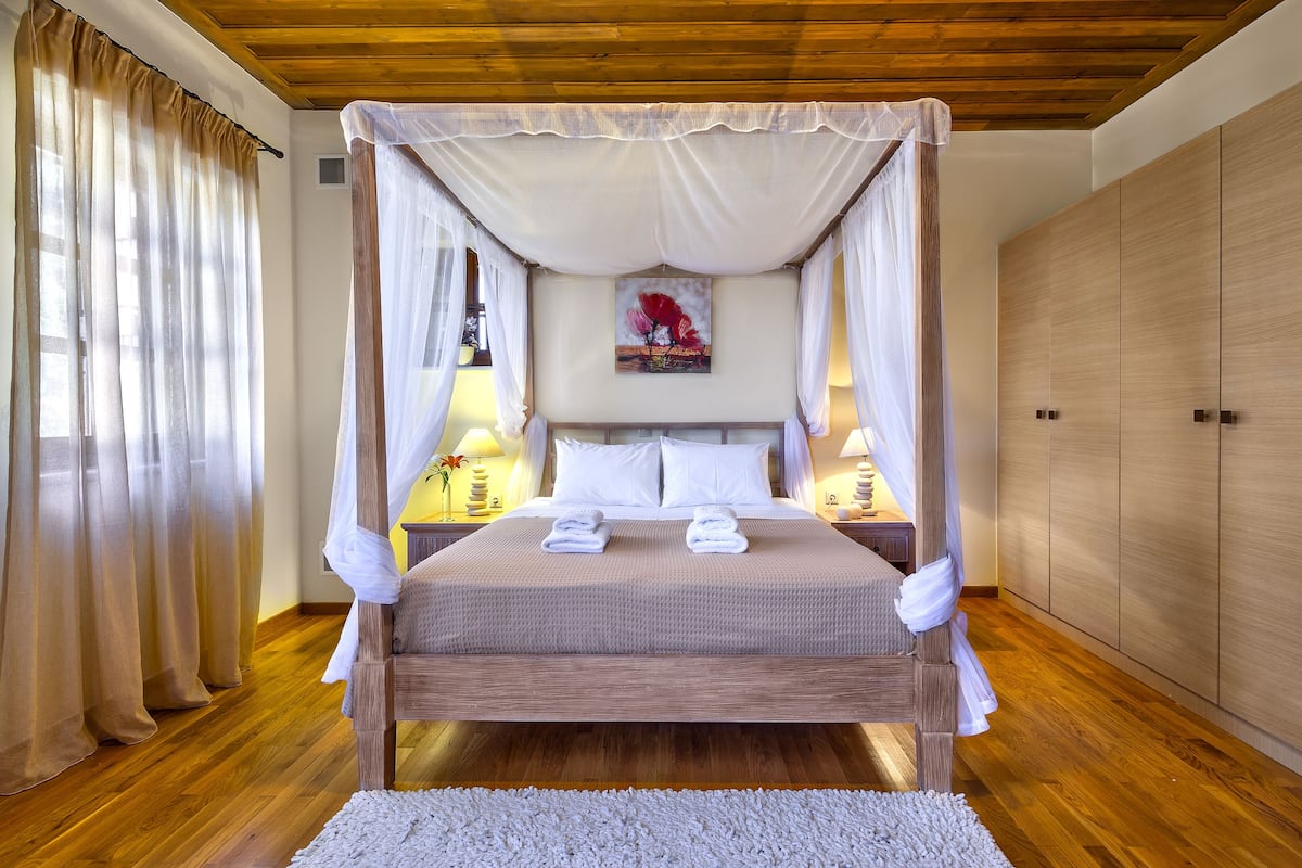 A four-poster canopy bed with white linens, two bedside lamps, and a painting on the wall.