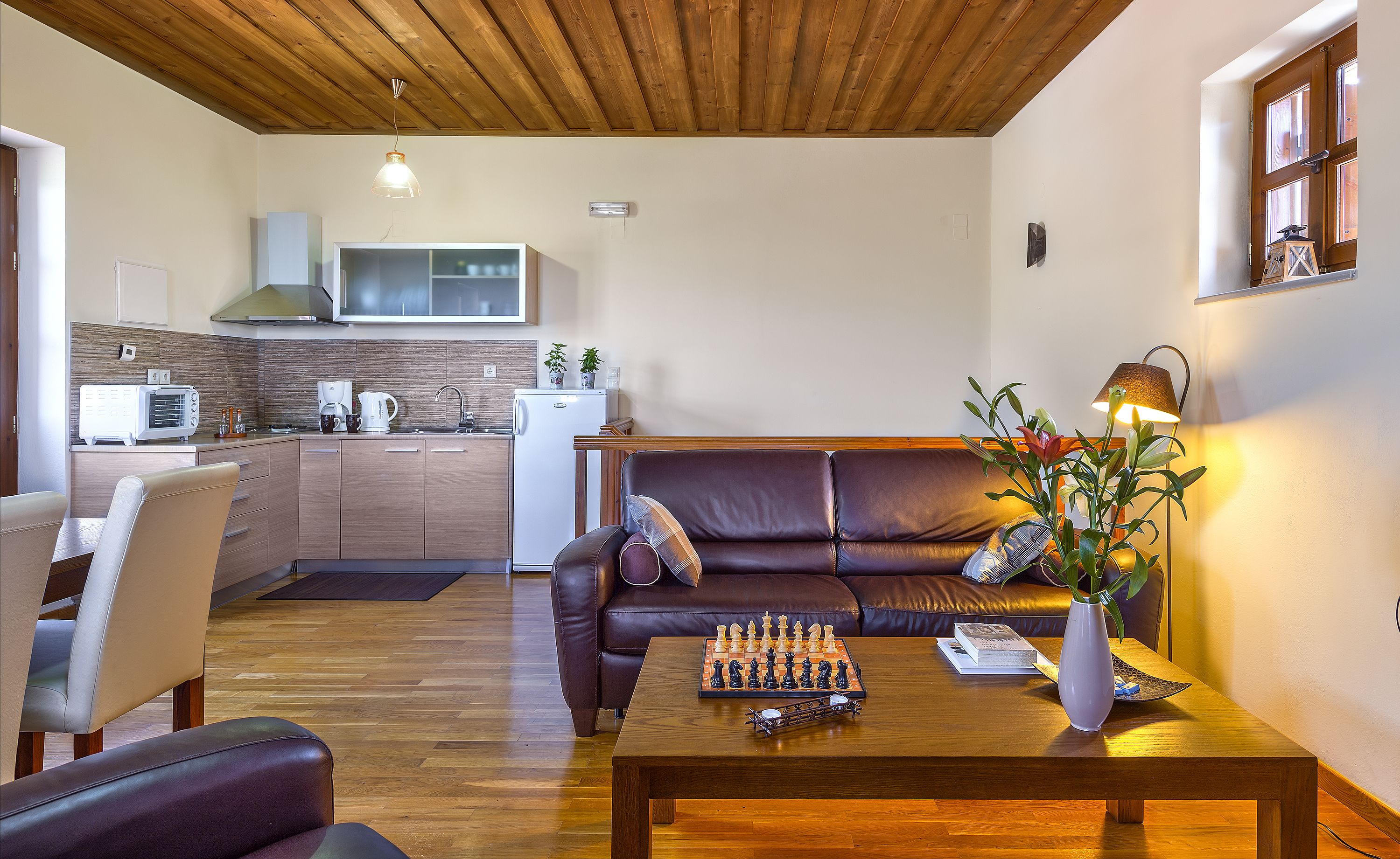 Un soggiorno moderno con soffitto in legno, un divano in pelle marrone, un tavolino da caffè con scacchi e una cucina sullo sfondo.