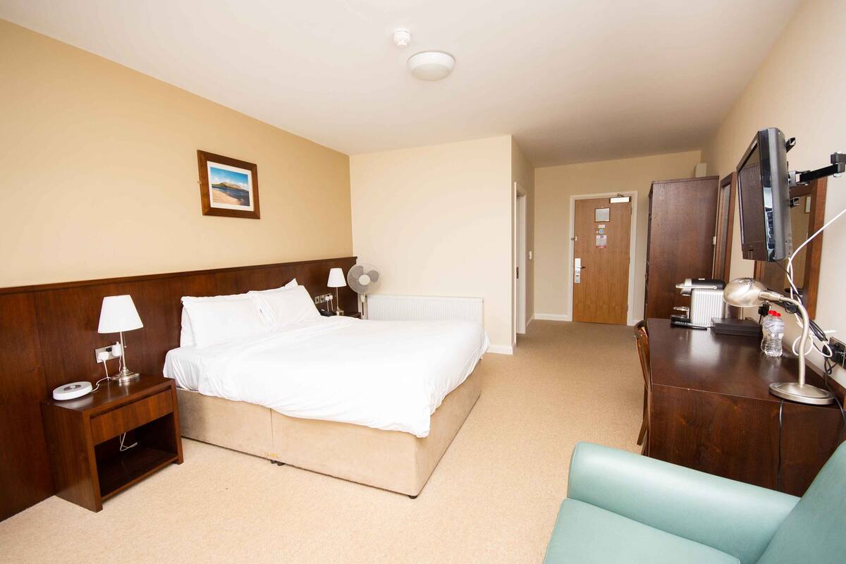 Quarto de hotel com cama, mesa de cabeceira, luminária e escrivaninha com espelho e luminária.