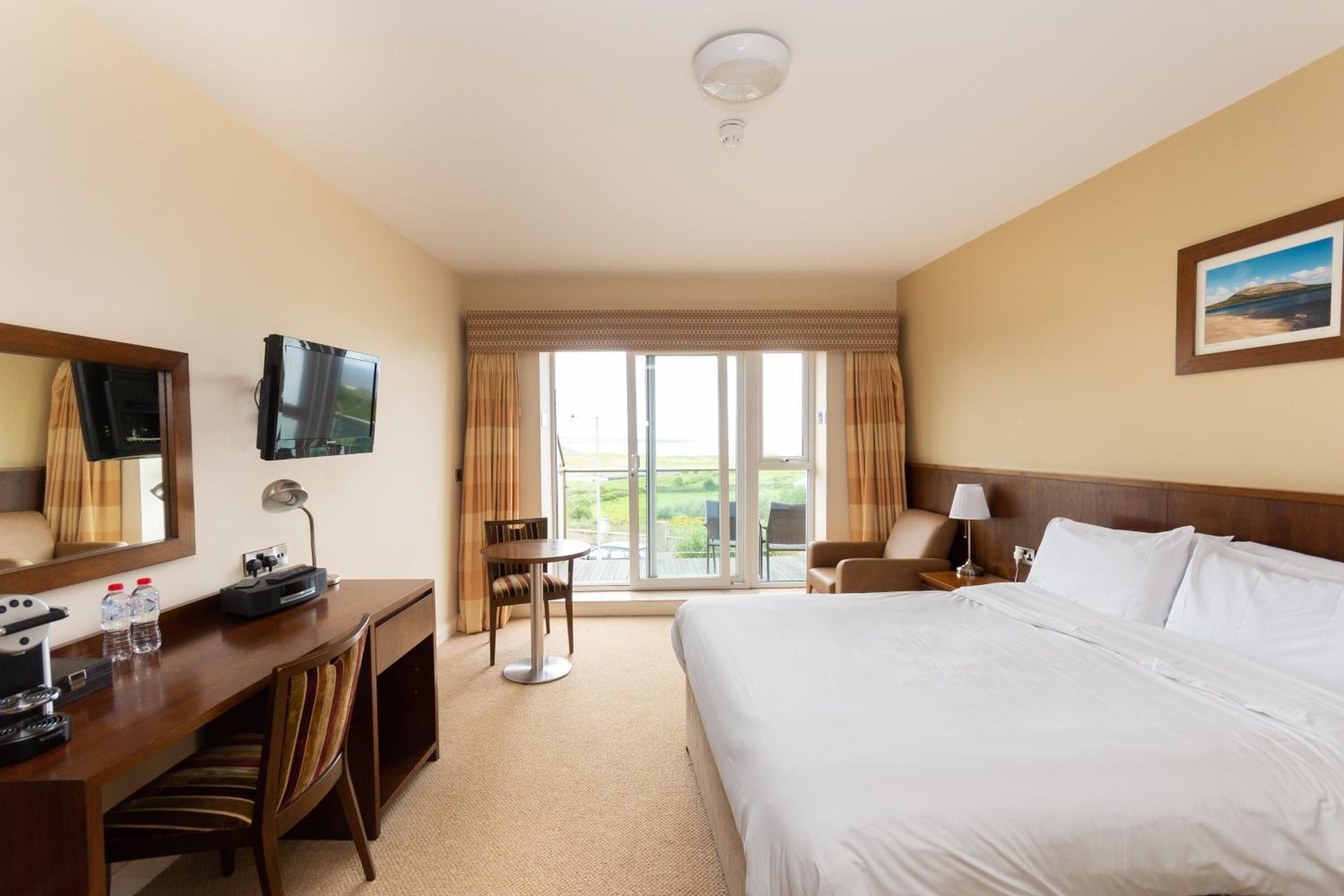 A hotel room with a large bed, a desk with a computer and phone, a chair, a TV, and a window with curtains.