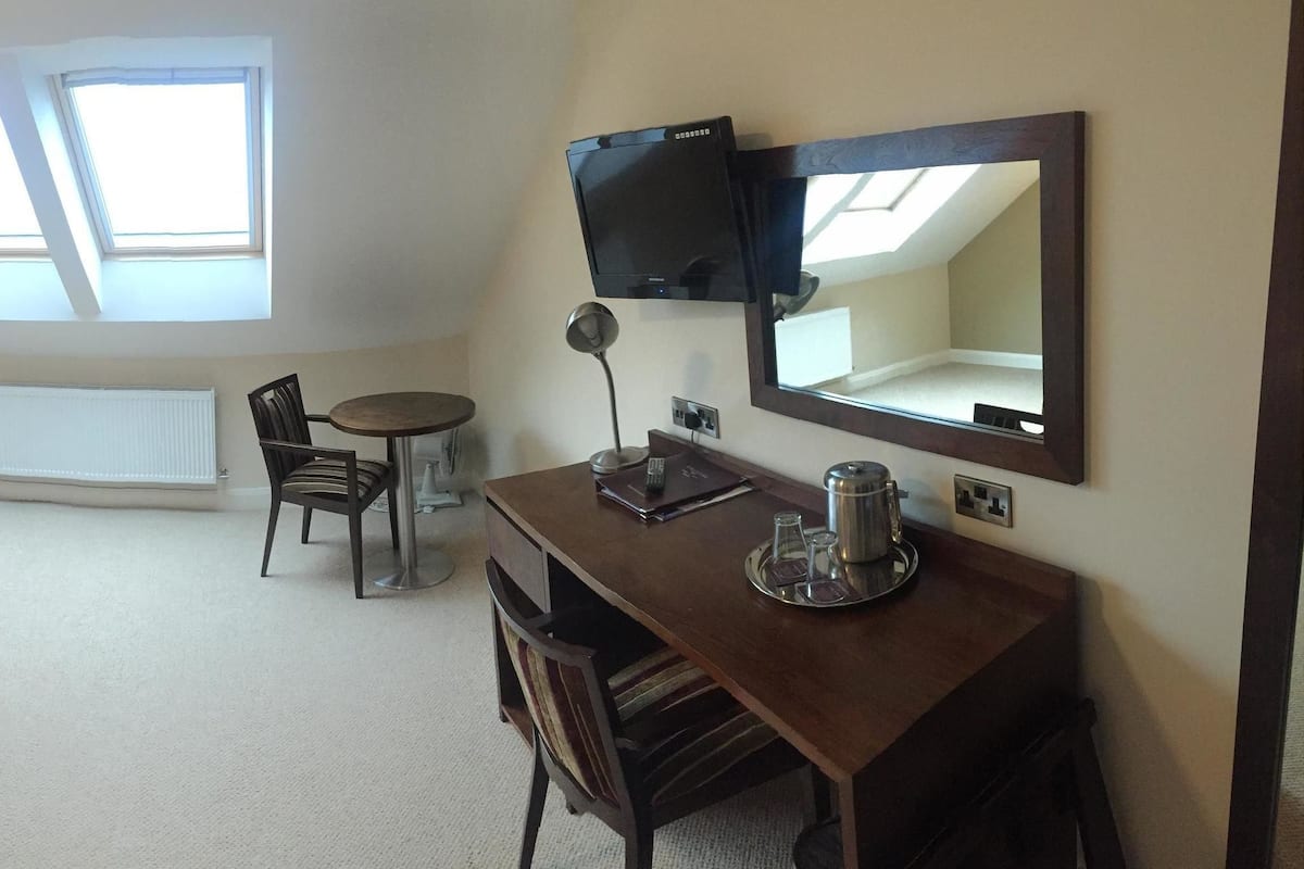 Quarto de hotel com cama, abajures, escrivaninha com cadeira, mesinha, televisão e janela com vista.