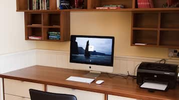 Un escritorio de madera con un monitor de computadora, teclado e impresora. Hay una silla negra con patas de metal. Encima del escritorio, hay estanterías de madera con libros y objetos decorativos.