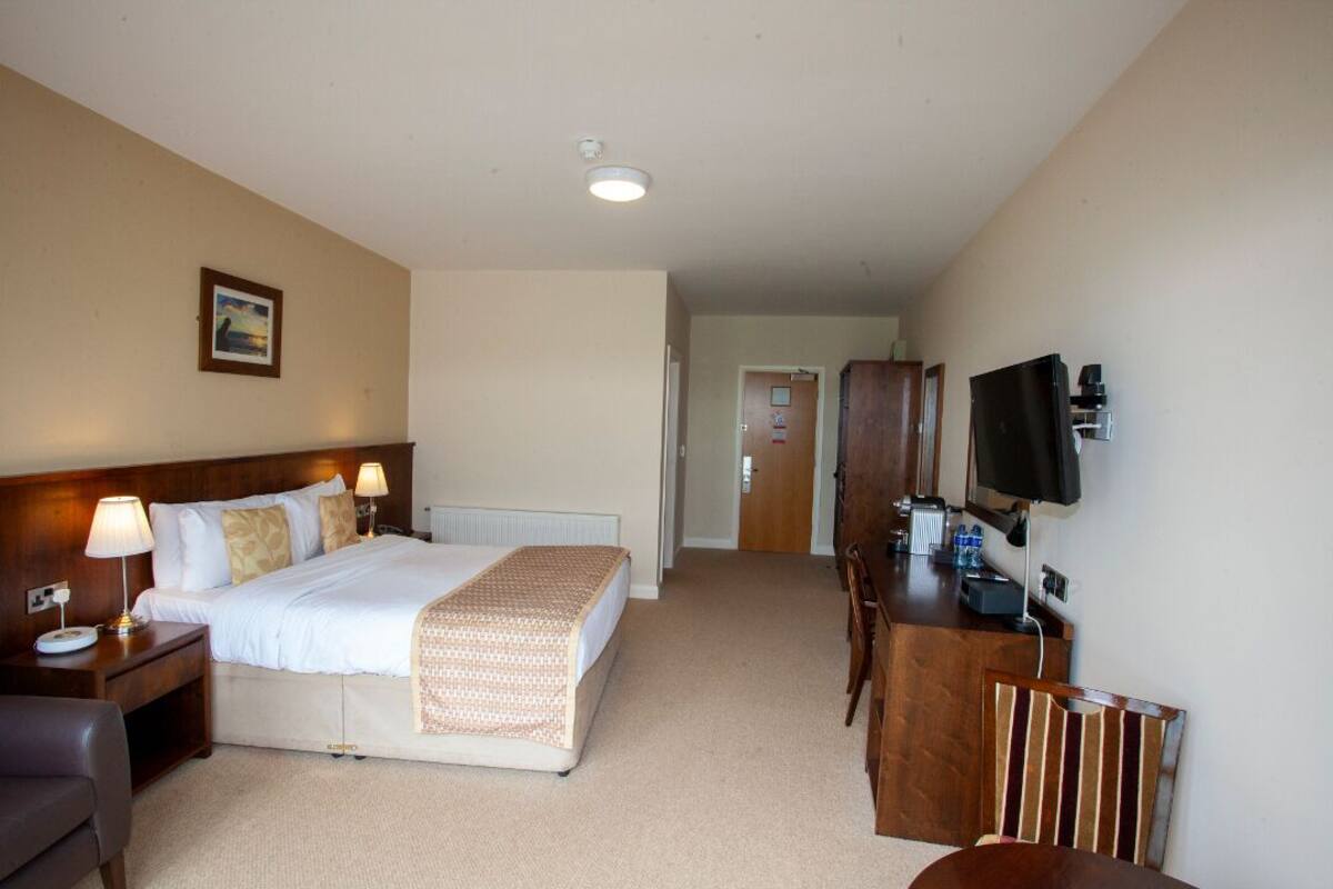 Quarto de hotel com cama, abajures, escrivaninha com cadeira, televisão e um quadro na parede.