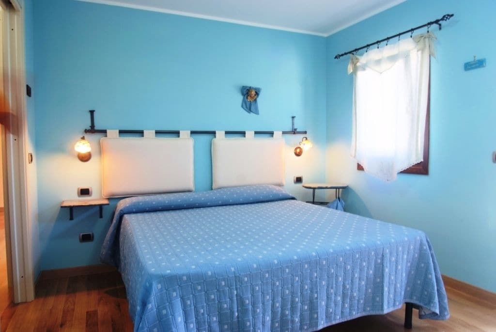 Un dormitorio con cama, dos mesitas de noche, una lámpara auxiliar y una ventana con cortina, además de luces fijadas a la pared.