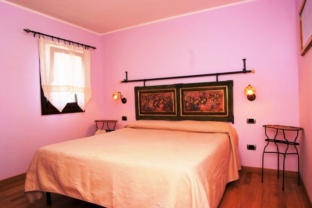 A bedroom with a bed, a window with curtains, two wall-mounted lamps, and a small table.