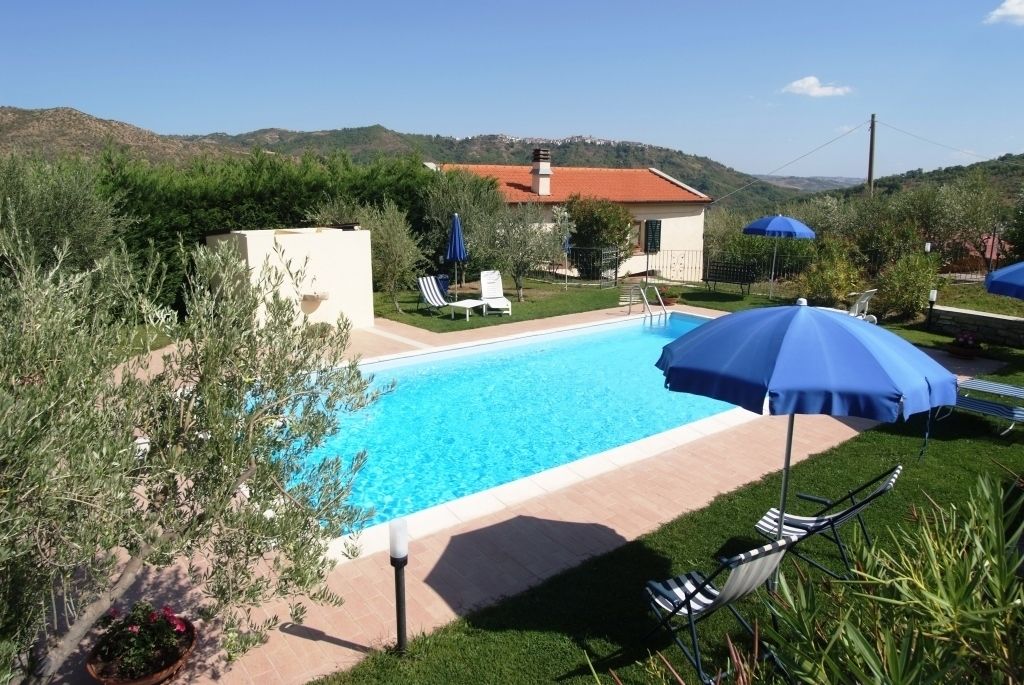 A swimming pool with lounge chairs and umbrellas, surrounded by greenery and hills in the background.