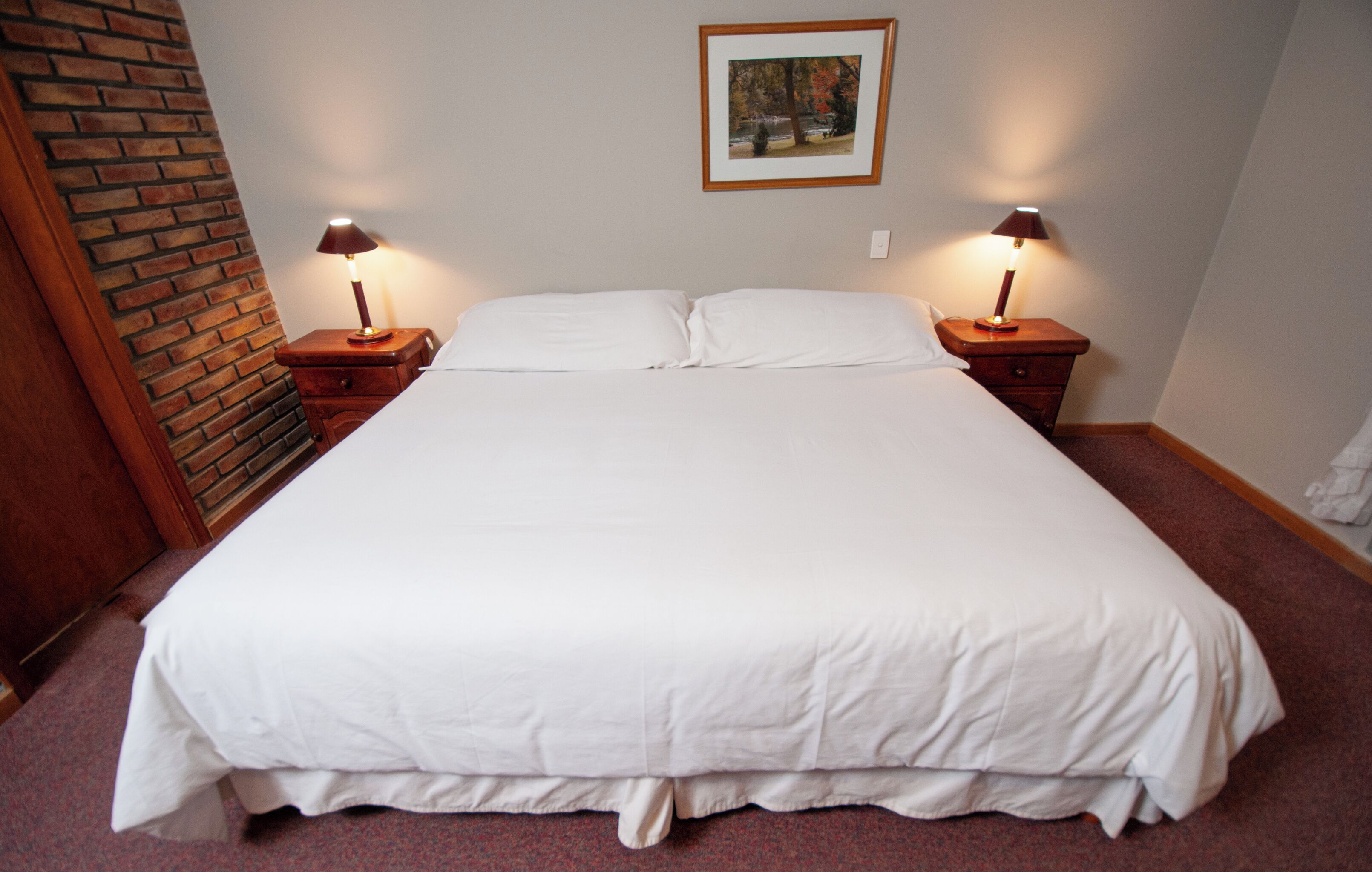 A neatly made bed with white linens, two bedside lamps, a framed picture on the wall, and a brick accent wall.