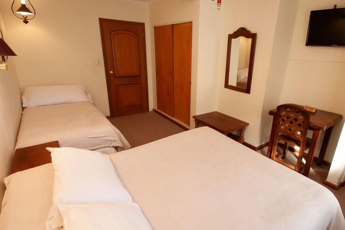 Ein Hotelzimmer mit zwei Einzelbetten, einem Holzschrank, einem kleinen Tisch, einem Sessel und einem Spiegel an der Wand.
