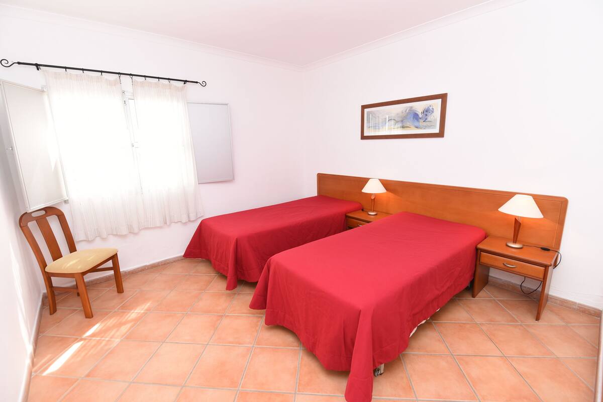 A room with two single beds, a chair, a nightstand with a lamp, a whiteboard, and a framed picture on the wall.