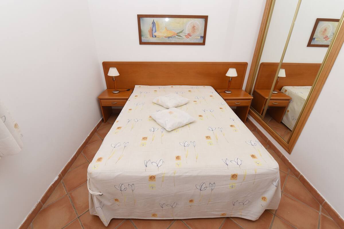 A neatly made bed with a floral bedspread, two bedside tables with lamps, a large mirror, and a painting on the wall.