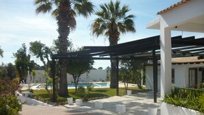 A covered outdoor area with a pool, palm trees, and a white building with a tiled roof. - Monte dos Avós Village - Pets Friendly (Tavira)