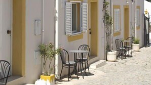 A building with white walls and yellow trim, featuring multiple doors and windows with shutters. There are outdoor tables and chairs, and potted plants are placed along the building's exterior. - Monte dos Avós Village - Pets Friendly (Tavira)
