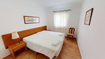 A hotel room with two beds, a nightstand, a chair, a window with curtains, and two framed pictures on the walls.