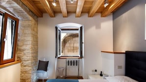 A bedroom with a stone wall, wooden ceiling beams, a bed with white and blue bedding, a clear chair, and a window with a view of an archway. - Villa Marta (Split)