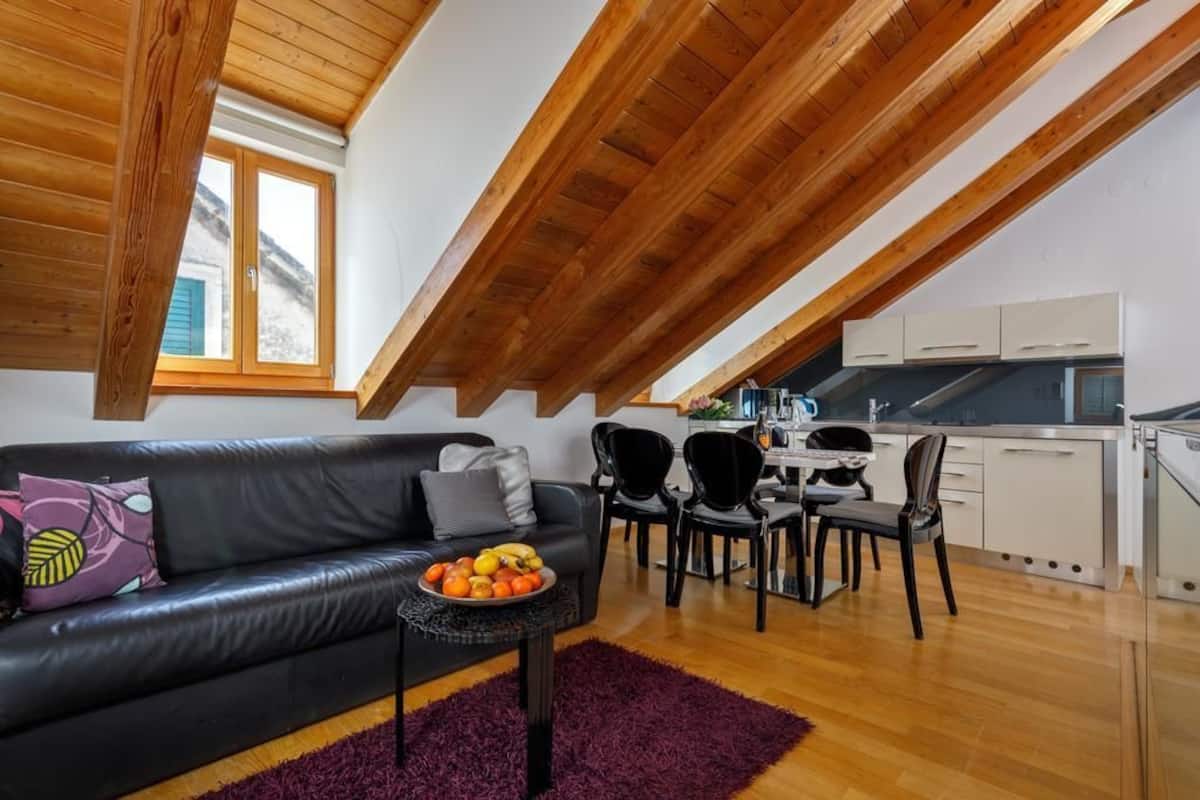 A modern loft apartment with a wooden ceiling, a black leather sofa, a dining table with chairs, and a kitchen area.