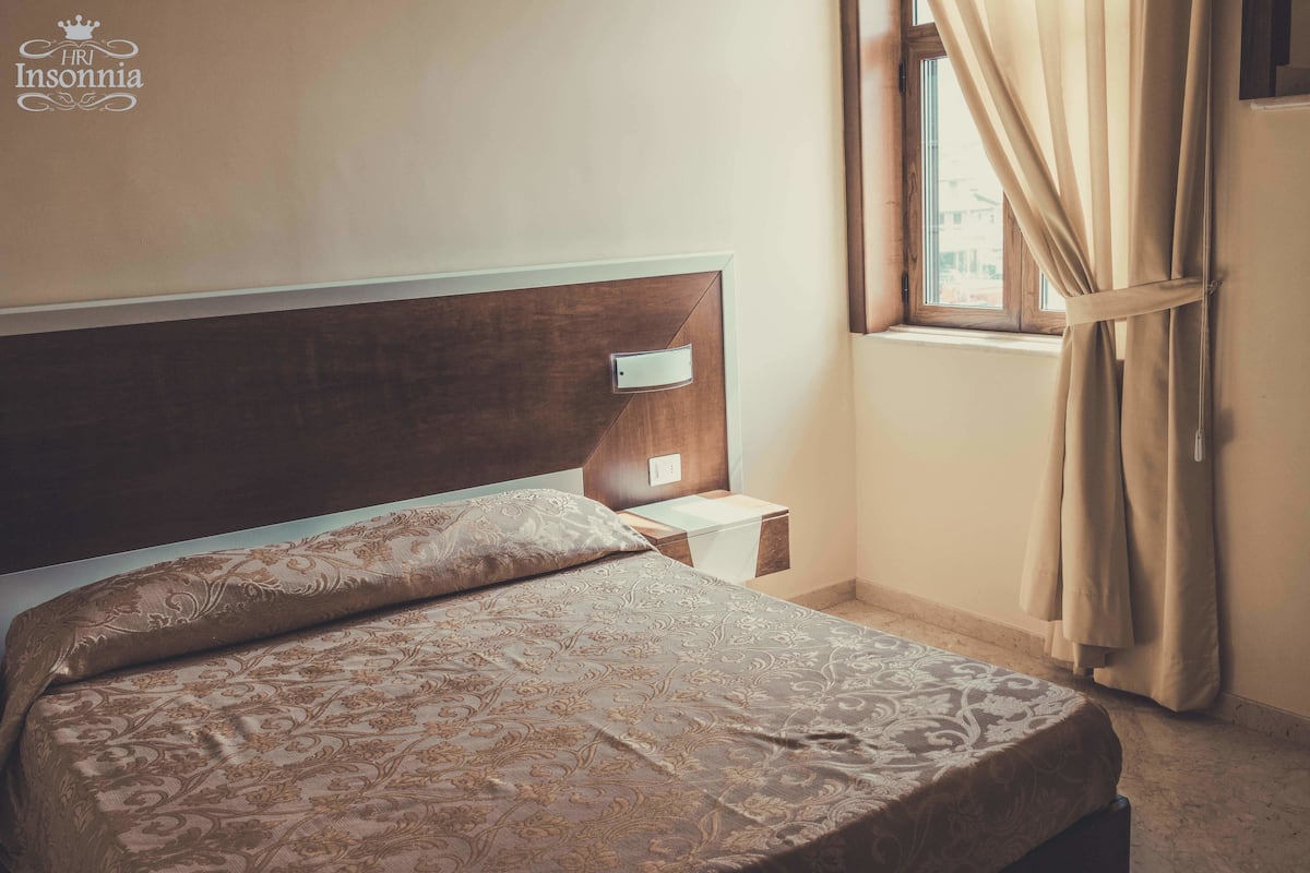 A neatly made bed with a patterned bedspread, a wooden headboard, a bedside table with a lamp, and a window with curtains.
