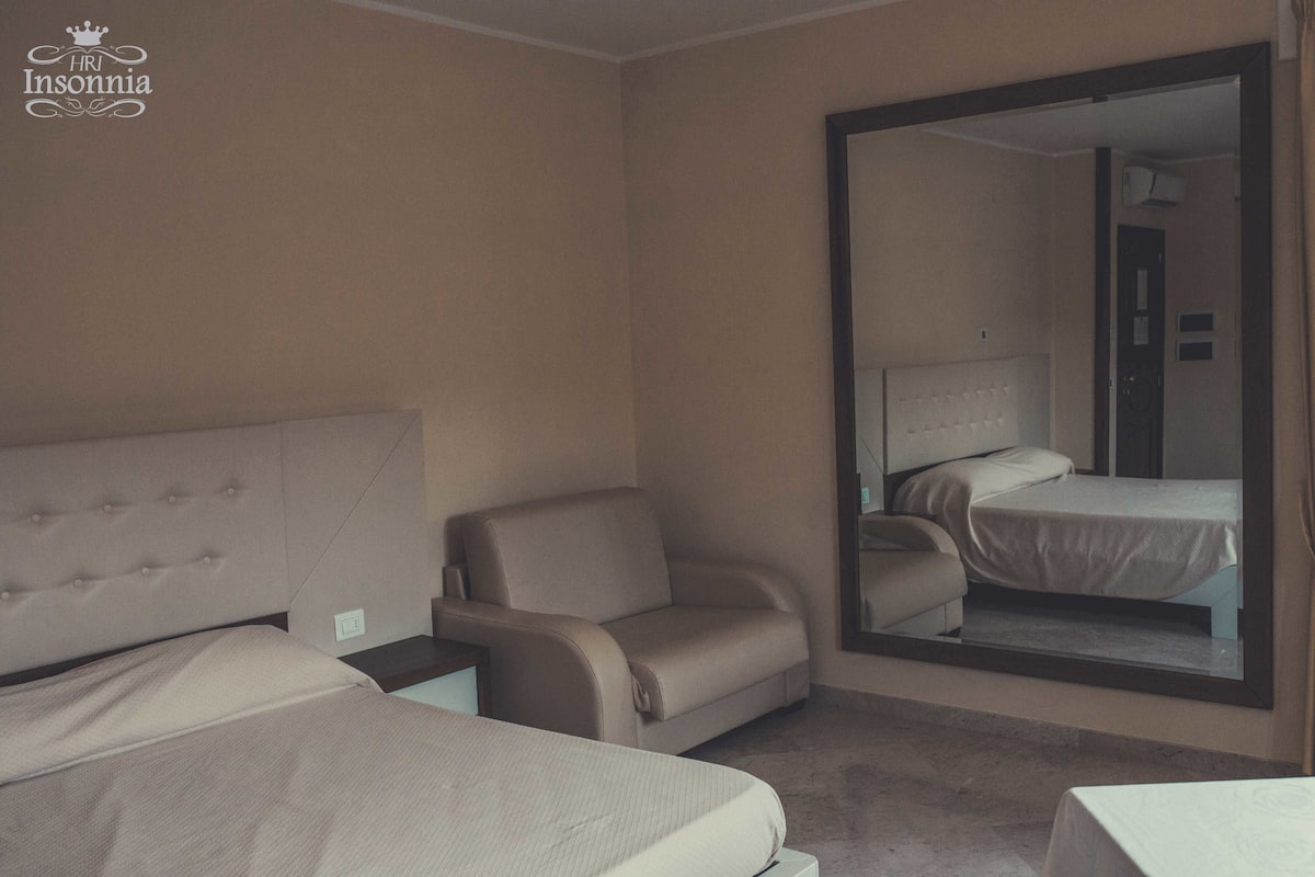 A hotel room with a large bed, a chair, a small table, and a mirror reflecting the bed.