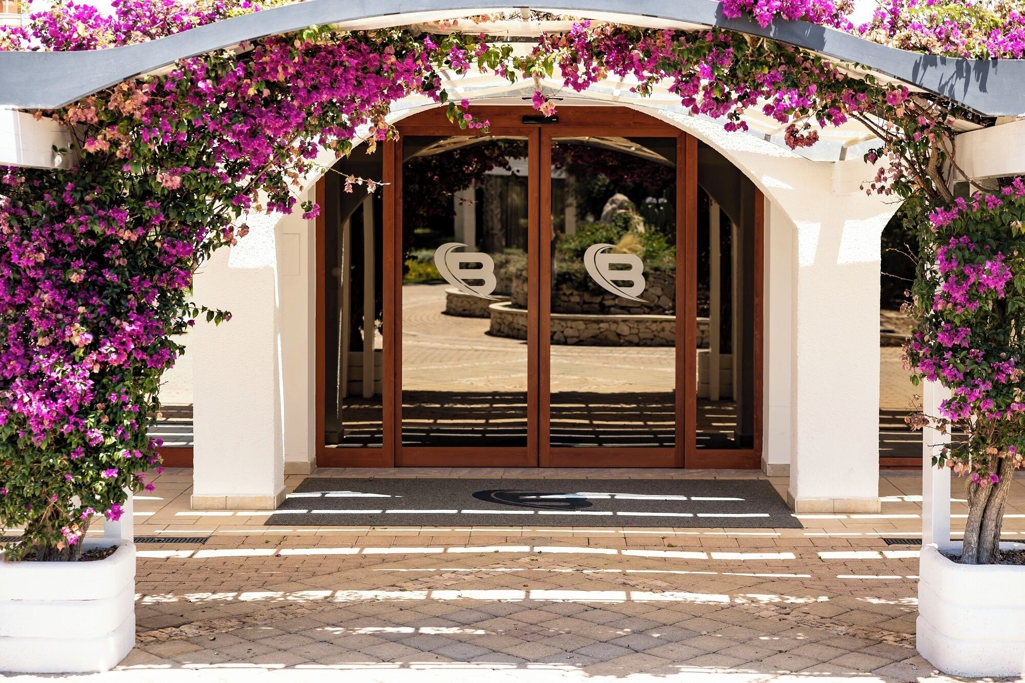 Entrada de hotel com porta de vidro, caminho de pedra e parede branca.